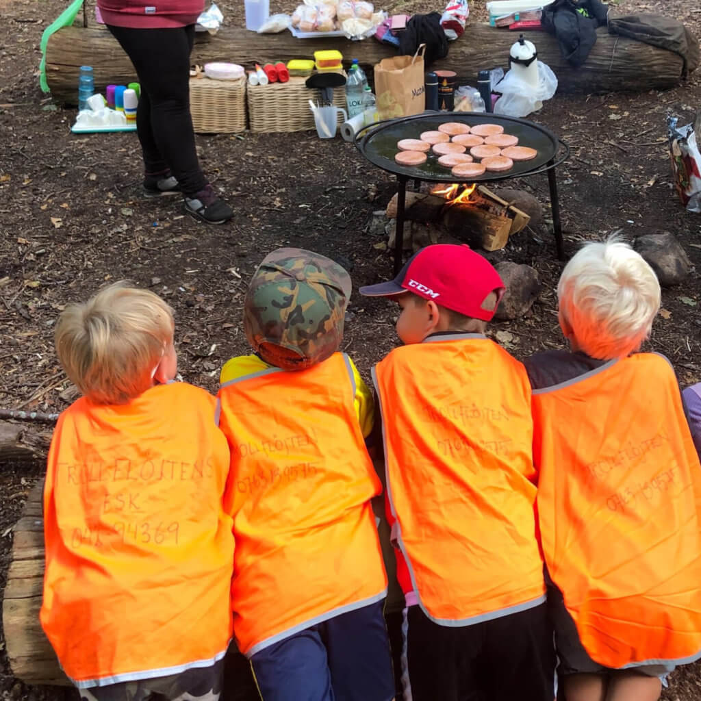Barn i orangea västar står på knä och väntar på mat vid stekbord utomhus i skogen